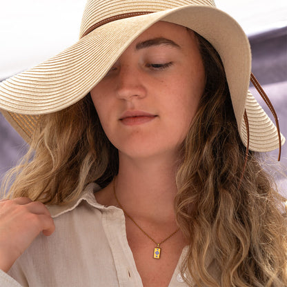 Woman wearing a wide-brimmed hat with a blurred background wearing a gold GAIA rectangular necklace