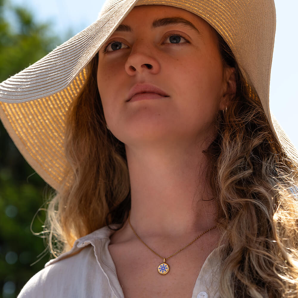 Woman wearing a wide-brimmed hat with a blurred natural background wearing a gold round SOLIS pendant nacklace with a purple sun