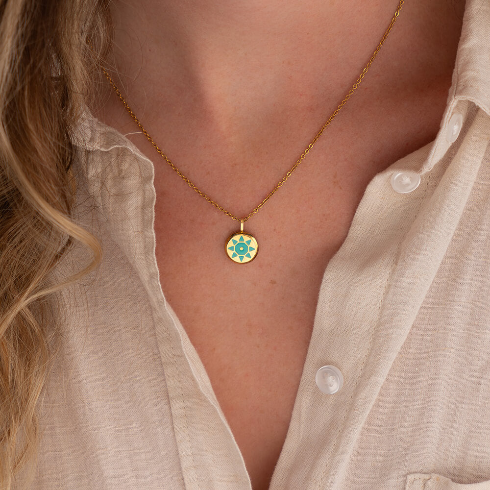 Gold SOLIS necklace with a round pendant with a sun design worn by a person in a light-colored shirt.