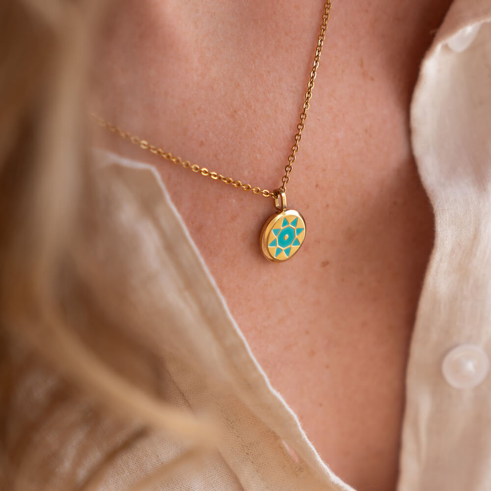 Gold SOLIS necklace with a gold and blue pendant on a person wearing a beige top.