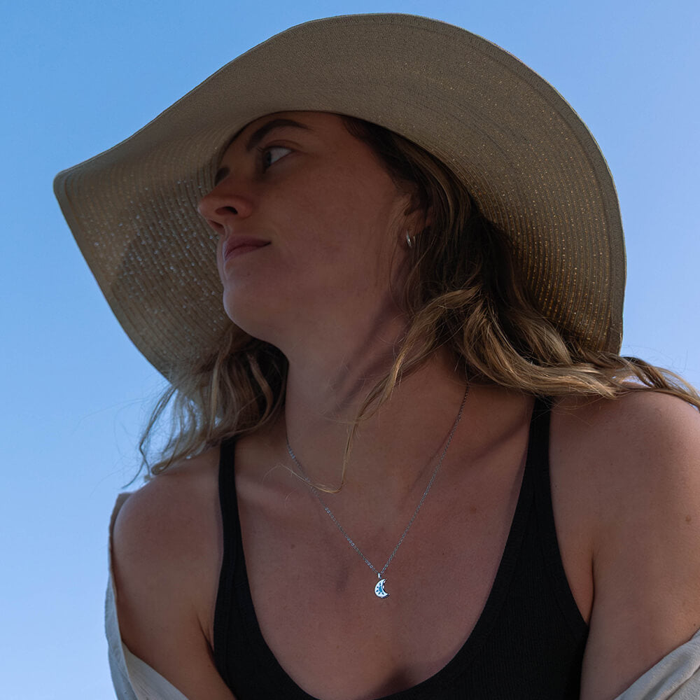 Woman wearing a wide-brimmed hat against a clear blue sky wearing a crescent shaped LUNA necklace