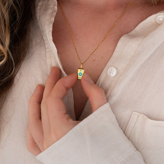 Person wearing a gold GAIA necklace with a blue pendant, close-up of the necklace and hand.